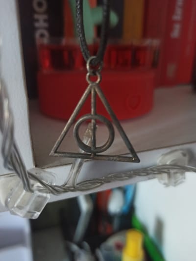 The image displays a close-up of a Deathly Hallows necklace suspended from a black cord. The necklace is made of a metallic material, possibly silver or pewter, and features the iconic triangle, circle, and line symbol. It is positioned in front of a blurred background that includes a red object, possibly a container, and some books or other items on shelves. The lower portion of the image shows a string of clear lights with wires, which appear to be Christmas lights or decorative lighting. The lighting is dim, and the focus is on the necklace. There is no indication of the city of Promyshlennaya, Russia, or any other specific location in this image.