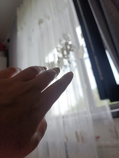 A close-up, low-angle shot shows a person's right hand, palm facing inward and fingers extended toward a sheer white curtain. The curtain is draped over a window with a dark curtain visible to the right. The background is blurred, but hints of greenery and light suggest an outdoor environment beyond the window. The scene appears to be indoors, near a window in what could be a residence in Novopokrovka, Kyrgyzstan. The lighting is soft and diffused, casting subtle shadows on the hand.
