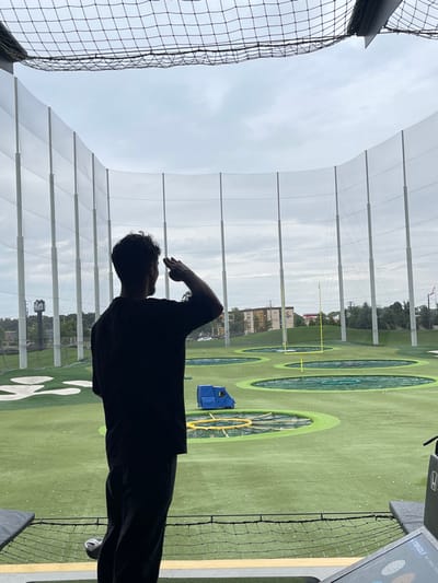 A male individual, seen from behind in profile, stands in a Topgolf hitting bay in Oxon Hill, United States, overlooking the driving range. He wears a dark short-sleeved shirt and dark pants, and has dark, curly hair. His right arm is raised, with his hand touching his forehead in a salute gesture, as he gazes towards the range. The environment consists of an artificial turf driving range with multiple circular target areas marked with green and yellow rings, scattered with numerous white golf balls. A blue utility vehicle is positioned on the turf, and a yellow goalpost is visible in the distance. Tall white poles support extensive black netting that encloses the range. Beyond the netting, a background of buildings, trees, and a road under an overcast sky is visible. A display panel partially visible on the right foreground shows "TOPGOLF" and a "HONDA" logo.