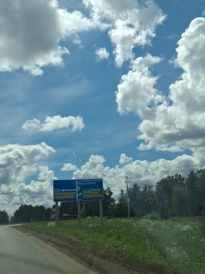 The image shows a roadside scene with a clear blue sky filled with scattered white clouds. In the foreground, a two-lane road curves to the left, bordered by a grassy area with wildflowers. Two blue road signs are prominently displayed on the right side of the road. The sign on the left indicates "M12 ЕКАТЕРИНБУРГ ПЛАТНАЯ ДОРОГА" (M12 YEKATERINBURG TOLL ROAD) with an arrow pointing left. The sign on the right indicates "M12 КАЗАНЬ ПЛАТНАЯ ДОРОГА 300 М" (M12 KAZAN TOLL ROAD 300 M) with an arrow pointing right. In the background, there is a line of trees and a utility pole. A white truck with a trailer is visible behind the signs. The scene appears to be a rural or semi-rural location, likely on a highway. The provided location of Уразгильды, Russia, suggests this is a view of the M12 highway near that settlement.