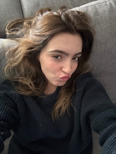 This image, taken in Nogueira e Silva Escura, Portugal, depicts a single female subject in an indoor environment. The individual, estimated to be a young adult, is positioned supine on a grey, textured upholstered surface, likely a sofa. She wears a dark, long-sleeved, knitted sweater with a prominent waffle-knit pattern. Her medium-brown hair, featuring lighter streaks, is loosely spread around her head. The subject faces the camera, maintaining direct eye contact, and displays a facial expression characterized by pursed lips. No other persons or significant objects are present in the visible portion of the scene. Illumination appears consistent with ambient indoor lighting.