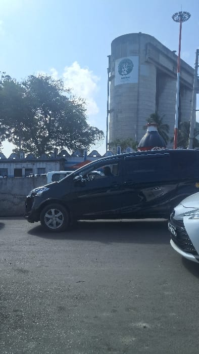 The image depicts a daylight street scene in the Ganjoni/Shimanzi ward of Mombasa, Kenya. The foreground consists of an asphalt road surface. In the midground, a dark-colored minivan, possibly black, is parked or stationary, oriented towards the left. The driver's side window is tinted, showing an indistinct dark silhouette of a potential occupant. To the right in the immediate foreground, the front bumper and part of the hood of a light-colored vehicle, likely white or silver, are visible, displaying a partially obscured Kenyan license plate.
A low, light-grey concrete wall with a distinctive crenellated top spans the midground from left to center, separating the road from an area behind it. Behind this wall, a white building with visible windows and other vehicles are partially obscured by a large tree with dense green foliage in the upper left.
The background is dominated by a massive, multi-sectioned light-grey concrete industrial building, consistent with a silo complex. A large circular white banner with a green logo and "Kuruwitu" text is prominently displayed on its side, identifying it as part of a Kuruwitu Cement facility. To the left of this industrial building, an oversized brown Coca-Cola bottle sculpture is visible. A tall, red and white striped high-mast light pole with a circular lighting array at its apex stands to the right of the industrial structure. Several palm trees are scattered throughout the background. The sky is clear and bright blue with sparse white clouds, indicating fair weather conditions. No specific individual actions or interactions are discernible.