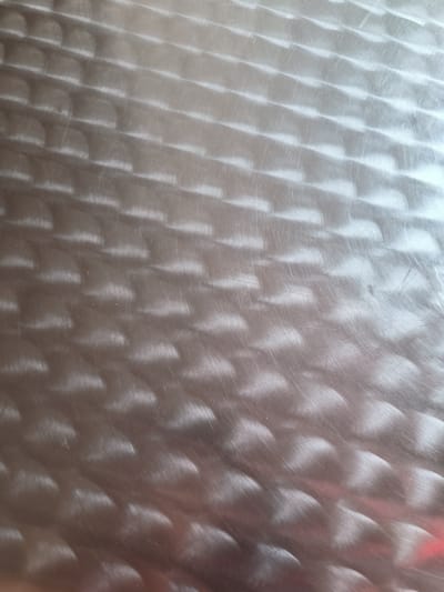 The image displays a close-up, overhead view of a textured metal surface. The surface has a brushed or patterned finish, creating a visual effect of repeating, elongated oval shapes or scales arranged in a diagonal grid. The metal appears to be silver or a similar metallic color, with areas of light reflection and shadow that highlight the texture. Fine scratches are visible across the surface, indicating wear or the material's age. The overall impression is one of a functional, industrial, or decorative metal material. There are no people, animals, or identifiable landmarks present in the image, and it is not possible to determine its specific location within Kladno, Czechia, based solely on the visual information.