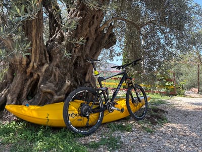 In Kanjoši, Montenegro, a yellow kayak is positioned horizontally on the ground, leaning against the base of a large, ancient olive tree. A black full-suspension mountain bike is placed on top of the kayak, with its front wheel resting near the bow of the kayak and its rear wheel extending towards the stern. The bike's frame is dark, and the wheels have white tire markings. The tree has a thick, gnarled trunk with exposed roots and is covered in green leaves, with branches arching over the kayak and bike. The surrounding environment consists of dry, rocky ground with patches of green grass and smaller plants. In the background, there are more olive trees, some vegetation, and a glimpse of hills or mountains under a clear blue sky. The scene appears to be an outdoor, rural setting.