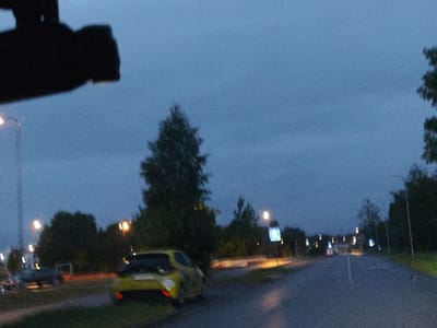The image captures a dimly lit outdoor scene, likely during dusk or evening, with a hazy, blue-grey sky. The foreground features a dark, silhouetted object, possibly a vehicle's interior or a dash-mounted device, obscuring the upper left portion of the frame. To the right of this obstruction, a street light with a warm yellow glow is visible.

Further into the scene, a yellow hatchback car is parked on the side of a road. The car is facing away from the camera and its taillights are illuminated. Beyond the car, a line of trees and bushes forms a dark backdrop. Several street lights are scattered along the road, some emitting a yellow light, while others are less distinct or obscured by foliage.

In the middle ground, there is a road with a white lane marker. The road appears to be wet, reflecting the light from the street lamps. Signs are visible along the roadside, one of which is a square blue sign with white symbols, mounted on a pole. The overall environment suggests a suburban or semi-rural setting within Tiskre, Estonia, given the presence of typical street infrastructure and vegetation. The image quality is somewhat grainy, consistent with low-light photography.