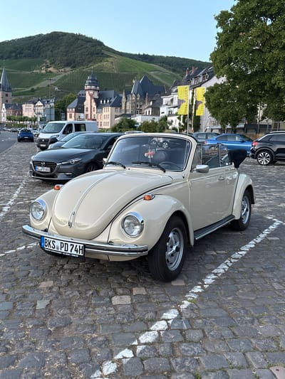 A beige Volkswagen Beetle convertible is parked on a cobblestone street in Bernkastel-Kues, Germany. The car is facing forward, with its front end towards the viewer. The convertible top is down, revealing the interior of the car. The license plate of the car reads "BKS PD 74H". In the background, there are several buildings, including a church with a tall spire. Vineyards cover the hills in the distance. Two yellow flags with black bears are hanging on a building on the right side of the image. Other cars are parked in the background, including a dark gray Mazda and a blue BMW. The sky is clear and blue.