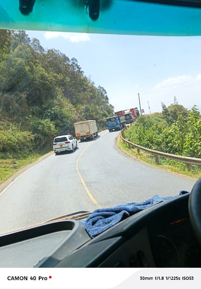 A winding asphalt road is seen from inside a vehicle. A white car follows a box truck. Further ahead, two trucks are visible, navigating the curve. Lush green vegetation covers the hillside on the left and right of the road. A metal guardrail lines the right side. Overhead, the sky is bright with a few clouds. Part of the vehicle's interior is in the foreground, including the dashboard.