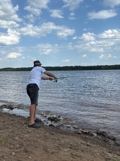 A man is fishing at the edge of a body of water. The man is wearing a white t-shirt, dark shorts, and a camouflage hat. He is holding a fishing rod with both hands and appears to be reeling in the line. The body of water extends into the distance, with a line of trees visible on the opposite shore. The sky is blue with scattered clouds. The ground under his feet is made up of dirt and small rocks. This image was likely taken in or near Doksha, Russia, given the request.