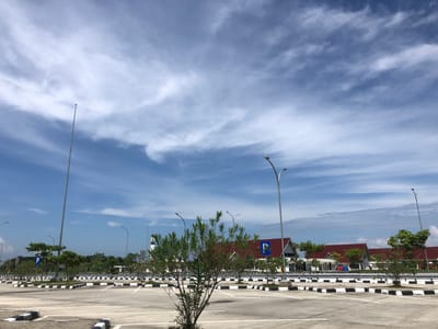 The image depicts an outdoor scene at what appears to be a parking area in Gohor Lama, Indonesia. The sky is bright blue with scattered white clouds. In the foreground, there is a paved parking lot with white and black striped parking bollards. Several streetlights are visible throughout the parking area. A few small trees are planted within the parking lot. In the middle ground, there are a series of modern buildings with red roofs, some with white walls. Beyond the buildings, a lush green treeline indicates a natural environment or parkland. A tall, slender tower, possibly a communication tower, stands prominently on the left side of the image. There are no people or vehicles visible in the scene. The overall impression is one of an open, well-maintained, and contemporary urban or semi-urban environment.