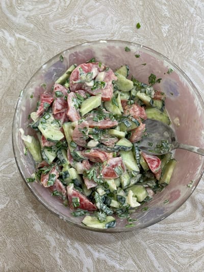 A close-up, overhead shot captures a clear glass bowl filled with a chopped salad. The salad consists of chopped tomatoes, cucumbers, and green onions, all coated in a creamy dressing. A silver spoon is partially submerged in the salad, suggesting it has been recently served or is about to be eaten. The bowl rests on a textured, beige tablecloth, which has a subtle, wavy pattern. The lighting is bright, indicating it's daytime, and there are no other people or objects visible in the frame. The setting appears to be a domestic environment, likely a kitchen or dining area in Kurskaya, Russia. The overall impression is one of a fresh, homemade salad ready to be enjoyed.