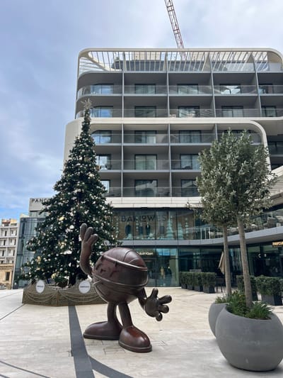 The image captures an outdoor plaza in Saint Julian's, Malta, featuring a blend of modern architecture and festive decorations under an overcast sky. In the foreground, a large, reddish-brown metallic sculpture of a stylized, round humanoid figure with three-fingered hands and large feet stands on a light-colored paved surface, with its right arm raised and left arm extended forward. A prominent dark linear stripe traverses the paving.
To the left of the sculpture, a tall, conical Christmas tree is heavily decorated with numerous white and gold spherical ornaments, topped by a multi-pointed star. Its base is encircled by a low, dark-colored barrier displaying "mercury" logos.
Behind the foreground elements, a multi-story building forms the midground. Its ground level features large glass storefronts, including one marked "BARLOWES" displaying mannequins in suits and decorative white Christmas trees, with "Calvin Klein" visible below. Another storefront to the right shows a "mercury" logo. To the right of the Christmas tree, two slender trees with light green foliage grow in large, round, grey concrete planters. On the far left, a multi-story building under construction with exposed concrete is partially visible.
The background is dominated by a modern, multi-story building with a light grey, curved facade, featuring numerous balconies with glass railings and individual sliding glass doors or windows. Some balconies show chairs. The top level of this building includes a distinctive, curved, light-colored architectural element. A red and white construction crane boom extends into the overcast sky above the right side of this structure. No individuals are present in the scene.