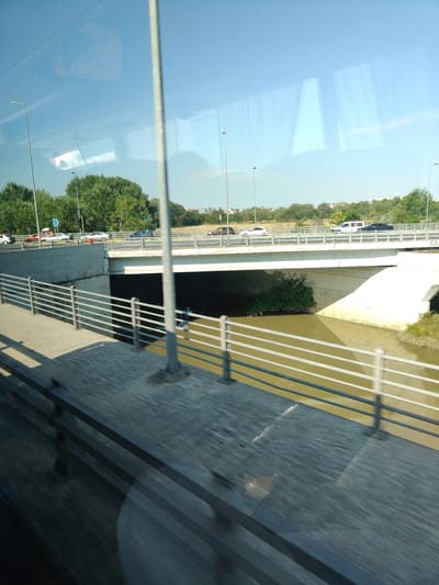 The image shows a highway overpass in Larissa, Greece, with traffic moving in both directions. The overpass spans a murky, brown body of water, likely a river or canal. The sides of the overpass are lined with white concrete barriers and metal railings. On the left side of the image, a pedestrian walkway runs parallel to the overpass, also with metal railings. Trees and vegetation line the banks of the water. The sky is clear and blue. Reflections of windows are visible on the surface of the water, indicating the presence of nearby buildings. The overall impression is one of urban infrastructure and daily life.