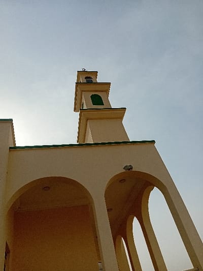 The image depicts a section of a mosque in Kariya, Nigeria, observed from a low-angle perspective looking upward. A cream-colored minaret, square-based, rises prominently against a pale, hazy sky, topped with a crescent moon finial. The minaret's central shaft features a green-framed arched window. The lower portion of the mosque building, also cream-colored, is visible with a series of arched openings and a green-trimmed roofline. Recessed lighting fixtures are present within the arched ceilings, and one external wall-mounted light fixture is affixed to the façade. The environment consists solely of the architectural structure set against the sky; no individuals, active subjects, or specific interactions are depicted.