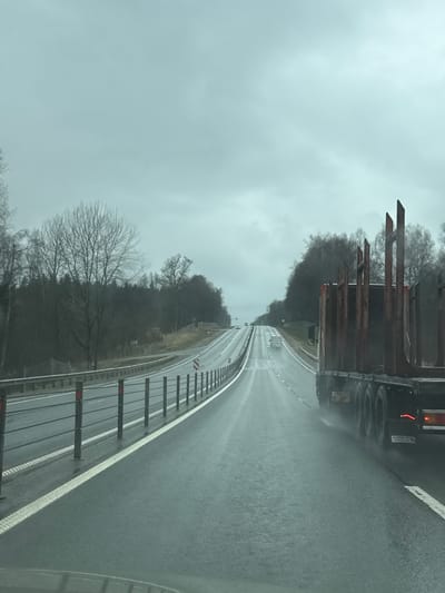 A view from a vehicle traveling on a highway under rainy conditions. To the right, a guardrail with white and red reflectors runs along the edge of the road. On the left side of the road, a vehicle, possibly a truck, is moving in the same direction. The sky is overcast and gray. Trees line both sides of the highway. There is no indication in the image that this is in Korobovo, Russia.
