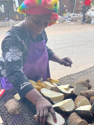 A person with a colorful headscarf and a purple apron is tending to a grill. The grill is covered with halved and quartered pieces of yams and plantains, which appear to be roasting or grilling. The person’s hands are visible as they handle the food on the grill. The setting appears to be outdoors, possibly a street scene, with a blurry background of buildings and debris.