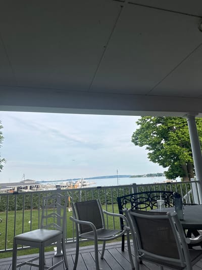 The image captures a view from a covered porch or deck overlooking a body of water, likely a harbor, in Harbor Springs, Michigan, United States of America. In the foreground, several outdoor chairs and a table are visible, suggesting a seating area. The chairs vary in design, with one white, ornate-backed chair and other more contemporary, lighter-colored chairs. A dark, patterned chair is also present. On the table, a clear plastic cup and a water bottle are placed.
Beyond the seating area, a metal railing separates the deck from a green lawn. In the middle ground, a line of docked boats, predominantly white, are visible along a waterfront with buildings in the background. The water extends to the horizon, where it meets a hazy sky with scattered clouds. To the right, a large, leafy green tree with a thick trunk is prominent, with a white pillar of the porch visible beside it. The overall scene depicts a relaxed outdoor setting with a view of a marina and a calm body of water.