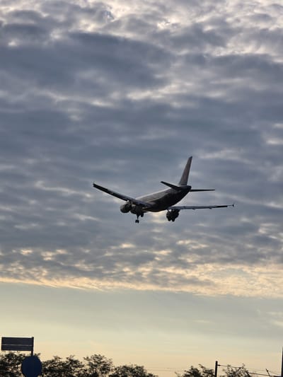 An airplane is depicted mid-flight against a cloudy sky. The plane, a passenger jet, appears to be in the process of landing or taking off, with its landing gear extended. The underside of the aircraft is visible, along with its wings and tail. The sky is filled with a mix of dark and light clouds, suggesting overcast conditions, possibly at dusk or dawn. In the lower portion of the image, silhouetted trees form a horizontal line, and a road sign is partially visible, indicating a location near Kelsterbach, Germany. The overall atmosphere is somewhat muted due to the lighting and cloud cover.