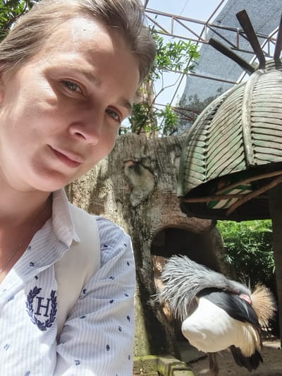 A woman is taking a selfie in what appears to be a zoo enclosure. She is wearing a light blue and white striped shirt with a crest on the left chest. Behind her, a crested bird is visible, with its head turned away from the camera and its crest feathers fanned out. The enclosure has a rocky background, greenery, and a man-made structure with a roof that resembles a mushroom or cap. The sky is visible through a mesh-like covering. The woman's face is partially obscured by the angle of the selfie, but her eyes are visible, and she has a slight smile. The overall impression is of a casual tourist visit to an animal exhibit in a sunny, outdoor environment.