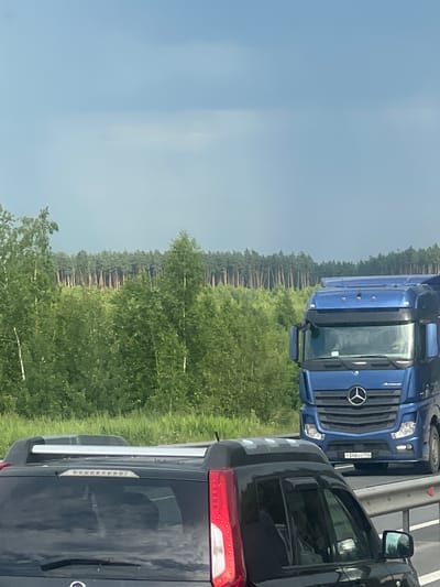 The image depicts a view from a vehicle traveling on a highway, likely in or near the city of Lukhovitsy, Russia. In the foreground, the rear of a dark-colored SUV is visible, featuring a roof rack. To the right of the SUV, a blue semi-truck is driving on the same highway. The truck has a license plate that reads "T 348 XO 750". Beyond the highway, a dense forest of green trees stretches across the landscape, with a line of taller, thinner trees forming a darker green band in the distance. The sky above is a muted blue with some overcast clouds. The overall scene suggests a daytime setting during the summer months.
