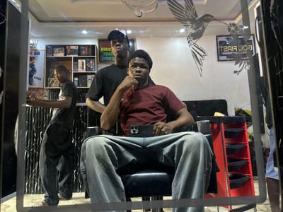 A medium, eye-level shot depicts three men inside a barbershop in Ogwashi-Uku, Nigeria, reflected in a streaked mirror. The primary focus is a man seated in a barber's chair, wearing a maroon t-shirt and light grey jeans, holding a smartphone. Behind him stands another man wearing a black t-shirt and baseball cap, with his hand placed on the seated man’s shoulder. To the left, a third man in a dark t-shirt and pants stands partially obscured by a black curtained partition.

The barbershop is decorated with product displays on shelves, and framed images are hung on the wall, including one with a stylized hummingbird and another with the words "Cut First." A red tiered barber's cart stands next to the seated man. The scene is lit by overhead lighting, including a decorative fixture hanging from the ceiling. The overall impression is of a candid moment captured in a working barbershop.