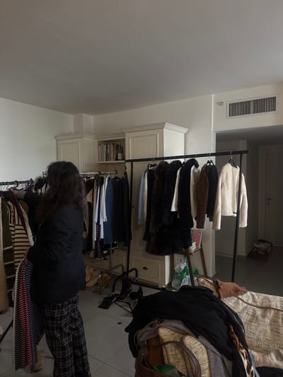 The image depicts an indoor scene within a room in Kfar Yona, Israel. One individual, viewed from behind, stands center-left, with long, dark, curly hair. They are wearing a dark blazer over a lighter top and dark plaid trousers, and are holding a striped garment (red, white, and blue) from a black metal clothing rack. This rack is laden with various garments, including striped tops, skirts, and trousers.
To the right, a second black metal clothing rack displays multiple coats and jackets, including dark faux fur-like items, a brown jacket, and a white sherpa-style jacket. In the background, white built-in cabinetry with decorative molding features an open shelf containing books on the left side, and closed cabinet doors on the right. The room has light-colored walls, a gray tiled floor, and a white ceiling. An air conditioning vent is visible on the upper right wall.
In the foreground, to the right, a light-colored, possibly upholstered, furniture piece is partially covered by dark and light clothing items, including a brown knitted top and a leopard print item. A brown leather handbag is also visible on this furniture piece. On the floor, near the right clothing rack, two plastic water bottles and a small wooden stool or stand are present. Light-colored shoes are on the floor near the left clothing rack. A doorway with a closed light-colored door is visible on the far right. A stack of folded items lies on the floor near this doorway. The overall scene suggests an environment for clothing organization, display, or selection.