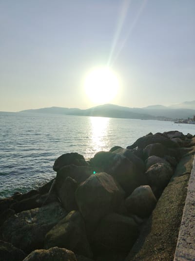 The image depicts a serene coastal scene at Marina di Casal Velino, Italy, likely during the late afternoon or early evening due to the low angle of the sun. The sun is a bright, prominent orb in the sky, casting a strong glare and creating a shimmering reflection on the calm, blue water. Rolling, hazy hills form the backdrop to the sea, with a hint of a town or village nestled along the coastline.
In the foreground, a dense collection of dark, rounded rocks forms a breakwater or seawall, providing a stark contrast to the softer tones of the sea and sky. Some of these rocks are covered in moss or lichen, indicating their exposure to the elements. To the right of the rocks, a textured, light-colored concrete structure, possibly a walkway or pier, runs along the edge of the image.
The overall atmosphere is tranquil and peaceful, evoking a sense of quiet beauty. The clear sky suggests fair weather, and the gentle waves imply a calm day. While the focus is on the natural landscape and the interplay of light and water, a few small figures can be seen in the distance near the shoreline, suggesting human presence and activity, perhaps enjoying the seaside. There is no visible text in the image.