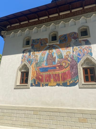 The image depicts a fresco on the exterior wall of a building, likely a church or monastery, located in Filioara, Romania. The fresco portrays a religious scene with numerous figures. In the center, a deceased figure lies on a raised bed covered with a red and gold patterned cloth, surrounded by other individuals who appear to be mourning or paying respects. Above them, celestial beings or angels are depicted. The fresco is rich in color and detail, with figures rendered in traditional Byzantine or Orthodox style. The building itself is white-plastered with ornate architectural details, including arched window frames with intricate carvings and a dark wooden roof with decorative trim. There are also two visible windows with dark wooden frames. The lower portion of the wall is constructed of light-colored brickwork. The sky above is a clear, bright blue, indicating it is a sunny day. The overall scene suggests a historical or religious context, typical of Orthodox Christian architecture found in Romania.