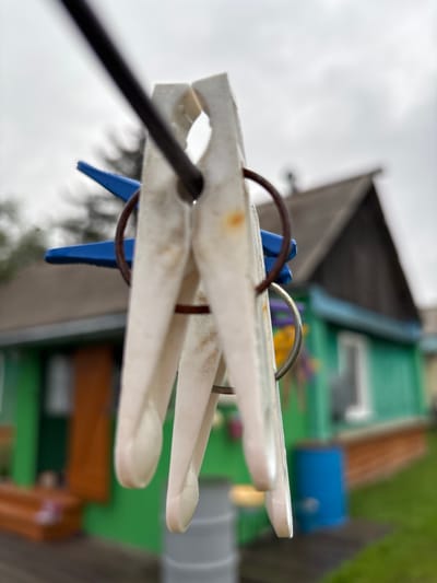 The image is a close-up, shallow depth of field shot of two white plastic clothespins on a dark clothesline. The clothespins are positioned vertically and appear to be slightly dirty, with some rust spots visible on one. Behind the clothespins, a blurry green house with a dark grey roof and a wooden porch is visible. A blue barrel and a grey cylinder are also present in the background. The sky is overcast and grey. The scene is set in an outdoor environment, likely a backyard, with green grass visible in the foreground and background. The location is specified as Gonja, Russia. No people or actions are depicted.