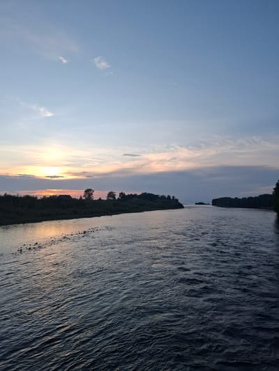 A wide, dark river flows towards a distant horizon under a twilight sky. The water is rippled, with small rocks visible along the near bank. A dense line of trees and a few buildings form a dark silhouette against the lighter sky on the left bank. The sky is a gradient of pale blue at the top to soft oranges and pinks near the horizon, with scattered clouds. The scene is tranquil and captures the essence of Vydrino, Russia, during the evening.