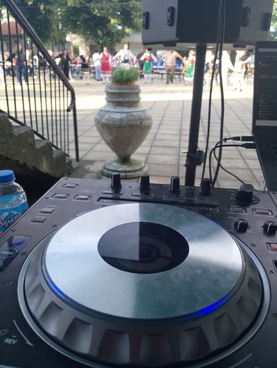 This image depicts an outdoor cultural event taking place in a paved courtyard or plaza, likely in Mogila, Bulgaria. In the foreground, a modern DJ setup is visible, featuring a black DJ controller with a prominent silver jog wheel, numerous buttons, and knobs. A laptop is connected to the controller on the right, and a black speaker on a stand is partially visible above. A plastic bottle of "Зелена" (Zelena) mineral water with a blue cap is positioned next to the controller on the left.
In the blurred background, a group of people is engaged in what appears to be a traditional folk dance. Many participants are dressed in vibrant traditional costumes, including long skirts, white blouses, and vests, holding hands in a line or circle, typical of a regional celebration or performance. Further back, trees and buildings suggest an open, community space. A large, ornate stone urn containing green plants stands in the mid-ground, adding a decorative element. A stone staircase with a black metal railing is visible on the left.
The lighting indicates it is daytime, likely late afternoon or early evening, with clear weather conditions. The scene captures a blend of traditional cultural expression with modern musical accompaniment. Visible text on the DJ controller includes "DECK", "REV", "GRID", "ADJUST", "SLIDE", "CLEAR", "FX", "JOG MODE", "NEEDLE SEARCH", "LOAD", "TAP", "FX MODE", "BACK", "VIEW", "JOG FEELING", "LIGHT", "HEAVY", "FAST", "KEY LOCK", and "TEMP".