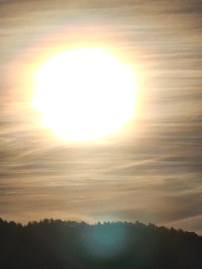 A bright sun dominates the image, surrounded by a hazy, streaked sky. The right side depicts a dark, tree-lined horizon. The colors are muted, with a predominantly warm and yellowish tone. Two faint, small dots appear in the sky to the lower-right of the sun. A bright lens flare appears on the right side of the image.