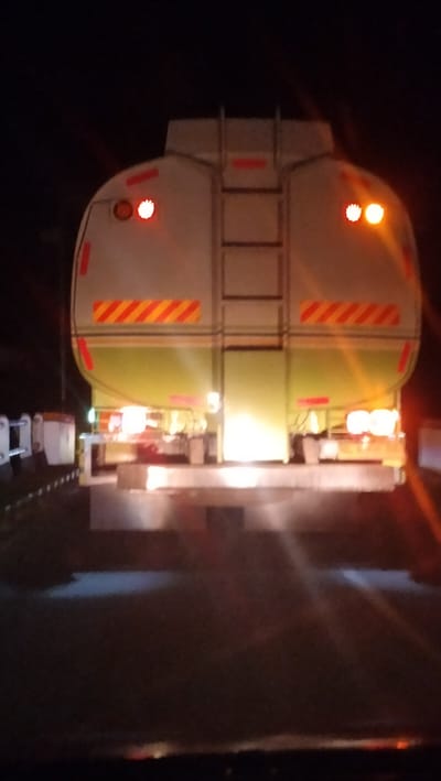 This night-time image, captured in Meucat Pangwa, Indonesia, presents the rear view of a large tanker truck, presumably from the perspective of a vehicle following it on a road. The scene is enveloped in darkness, characteristic of night, with illumination primarily originating from the truck's lights.
The tanker truck features a cylindrical tank, which is light gray or white on its upper portion and transitions to a pale green or yellow color on its lower half. A sturdy metal ladder is visible running vertically up the center of the tank's rear. For enhanced safety and visibility, the truck is equipped with several reflective markings: solid red strips are placed horizontally and vertically, alongside prominent red and yellow diagonal striped patterns positioned on both sides of the tank.
Various lights are active on the truck's rear. Near the top, there are two pairs of circular lights: an inner set of red lights and an outer set of amber or orange lights. At the bottom of the truck's rear, multiple bright white and yellow lights are illuminated, likely serving as taillights, brake lights, and possibly additional auxiliary lights, casting a strong glare. The rear bumper or underride guard is also reflective and brightly lit. To the left side of the truck, faint outlines of white road barriers or markers can be seen. No individuals are present in the image, and no legible text is visible on the truck or in the surrounding environment.