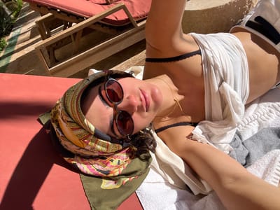 An individual, appearing to be a young woman, is shown in a close-up, upward-angled selfie, lying on her back on a red sun lounger cushion. She is positioned outdoors in Rissani, Morocco, under bright sunlight. Her head is wrapped in an olive green silk scarf with yellow, red, and pink patterns. She wears dark, round-framed sunglasses with reddish lenses and a delicate gold chain necklace. Her attire includes a black lace bralette under a loose white t-shirt that is partially lifted, exposing her midriff. A white terrycloth towel is visible beside her right arm (viewer's left). In the background, two additional wooden sun loungers with red cushions are present on a light-colored paved or tiled surface, with distinct shadows indicating strong illumination.