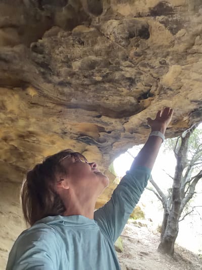 A person, viewed from below, looks up into a rock overhang, extending their right arm upwards towards the textured surface of the rock. The person is wearing glasses and a light blue long-sleeved shirt. A white wristband is visible on their right wrist. The rock overhang appears weathered and irregular. Beyond the overhang, a bright, sunlit area is visible, with a tree and sparse vegetation. The ground beneath the overhang is rocky and covered with leaves. The setting appears to be outdoors, possibly in a natural park or canyon area.