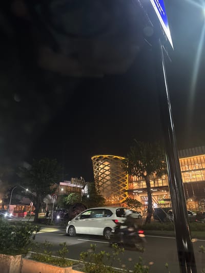 At night in Gunung Putri, Indonesia, a white car moves along a road lined with trees and streetlights. A building with illuminated windows is visible in the background. There is a cylindrical structure with a lattice design lit up. The photo is taken from an obscured perspective, possibly behind a sign, with a blurred object in the foreground.