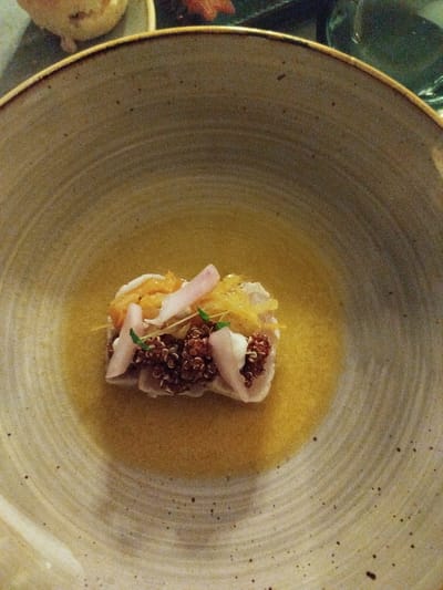 This is a medium-angle, close-up shot of a small, artfully presented dish of food in a large, shallow, speckled ceramic bowl. The dish is centrally located within the bowl and sits in a pool of light yellow liquid, likely a broth or sauce. The food itself is a small, rectangular mound comprised of layers of different ingredients. The bottom layer appears to be dark red quinoa. Above this, there are pieces of white fish and some yellow, shredded ingredients, possibly ginger or citrus zest. Thin, pale pink slices of what could be radish or thinly cut fruit are arranged on top, along with a few delicate green sprigs. A thin skewer or toothpick is partially visible, holding the ingredients together. The bowl has a textured, slightly rippled surface and a darker rim. The background is softly blurred, but hints of other dishes or tableware and a dark green surface are visible, suggesting a restaurant setting. The lighting is warm and intimate, possibly from overhead restaurant lights. The focus is sharply on the food, highlighting its delicate presentation. No people or text are visible in the image. The setting appears to be indoors, likely a dining establishment in Kendhoo, Maldives.