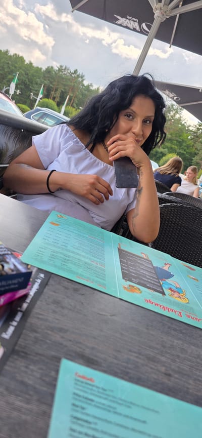 A woman with dark, curly hair and a black choker necklace sits at an outdoor table at a restaurant in Wietzendorf, Germany. She wears an off-the-shoulder white top and holds a smartphone to her face. A menu with a cartoon image of a blonde girl in a blue dress is open on the table in front of her. Behind her, two other women are visible at a separate table. The background includes trees and a cloudy sky.