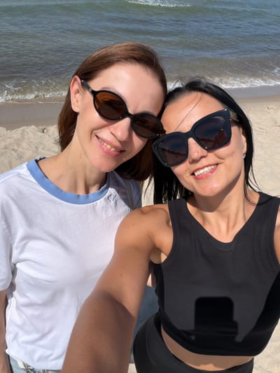 Two women are taking a selfie on a sandy beach with the ocean in the background. The woman on the left has brown hair and is wearing a white t-shirt with a blue collar. She is smiling and wearing dark sunglasses. The woman on the right has black hair and is wearing a black tank top. She is also smiling, wearing dark sunglasses, and has freckles on her face. The environment is sunny, with calm waves visible in the sea and a sandy beach. The image appears to be taken in Rybachy, Russia, given the context.