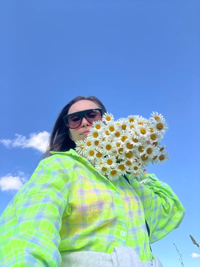 A low-angle, medium shot captures a person holding a bouquet of daisies against a bright blue sky with scattered white clouds. The individual, wearing dark-rimmed sunglasses and a neon green and blue plaid shirt, is positioned in the lower half of the frame. The flowers partially obscure their face. The background consists entirely of the sky and a hint of dry grass in the lower right corner. The location is identified as Pigozino, Russia.