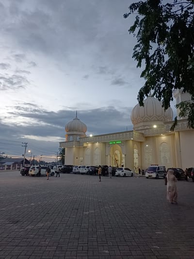 This is a photograph of the Baitul Huda mosque in Mee Tanjong Usi, Indonesia, taken at dusk. The mosque, with its distinctive dome and intricate architectural details, is illuminated by soft lighting. A parking lot in front of the mosque is filled with various cars. Several people are visible in the scene, some walking across the paved lot, while others are near the vehicles. The sky above is filled with dramatic clouds, hinting at the end of the day. A tree with lush foliage partially frames the right side of the image.