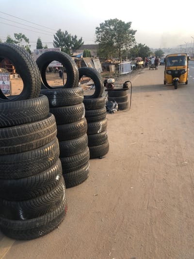 A stack of used tires sits on a dusty roadside, likely for sale. A yellow tuk-tuk travels along the road in the distance. Other vehicles and people are visible in the background. An air compressor and a few tires are positioned further away from the larger pile. The scene seems to depict a tire shop or vendor operating on the side of a busy street.