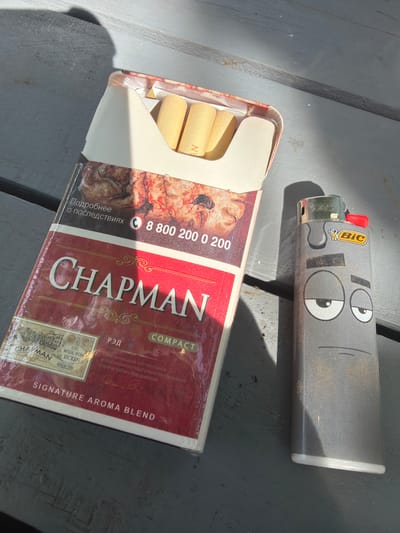 A BIC lighter with a cartoon face sticker on it rests on a dark gray wooden surface next to an open pack of Chapman Signature Aroma Blend cigarettes. The lighter is gray with a red button and a yellow BIC logo. The sticker features two large, wide-open eyes and a straight line mouth. The cigarette pack is mostly red with white and cream accents. The branding "CHAPMAN" is prominently displayed in large white lettering with ornate gold embellishments. Text on the pack includes "COMPACT," "SIGNATURE AROMA BLEND," "РЭД," and information about the tobacco blend and ingredients in Russian. A phone number, "8 800 2000 200," and the phrase "Подробнее о последствиях" (More about the consequences) are also visible. The cigarettes inside the pack have light yellow filters. The scene appears to be outdoors, possibly in a park or on a patio, with dappled sunlight casting shadows on the wooden surface. The lighting suggests it is daytime.