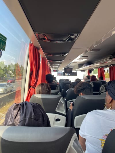 The image provides an interior view looking forward within a passenger bus or coach, which is in motion on a multi-lane highway. Specific geographical markers to pinpoint a precise location within Aulnay-sous-Bois, France, are not visibly present in the frame; however, the scene is consistent with a major road environment.

**Scene Composition:**
The perspective is from a passenger seat on the left side of the bus, angled slightly towards the right aisle. The bus interior features rows of modern, dark gray and black seating. Red curtains are drawn back at the windows along the right side of the vehicle. Overhead, the ceiling consists of dark gray panels interspersed with white sections, featuring integrated lighting and ventilation units with chrome accents. Overhead luggage compartments are visible. A small, dark display screen is centrally located over the aisle further down the bus. A green and white emergency exit instruction sign is partially visible on the window frame near the front of the bus on the left side.

**People:**
Approximately 15 individuals are visible from behind, occupying various seats within the bus. All are seated and facing forward. In the immediate foreground on the left, a person wears a dark, possibly navy or black, shiny head covering. To their right, closer to the camera, a person wearing a white t-shirt with a colorful graphic on the lower back (depicting yellow lightning bolts and other abstract shapes) and large gold hoop earrings is visible. Further into the bus on the left side, a person with dark, curly hair is observed holding a smartphone in their right hand, seemingly interacting with the device. Other passengers further down the aisle exhibit various hair colors, including dark brown, reddish, and lighter shades. No direct interactions between passengers are discernible.

**Outside Environment:**
Through the large windows on the right side of the bus, a multi-lane highway is visible. Traffic is moving in the same direction as the bus. At least two cars are in view: a light-colored (possibly silver or champagne) sedan in the lane immediately adjacent to the bus, and a red sedan further ahead in an adjacent lane. The road is bordered by green trees and other vegetation. The sky is clear and blue, indicating daytime conditions. Bright sunlight illuminates the scene, casting shadows within the bus interior.