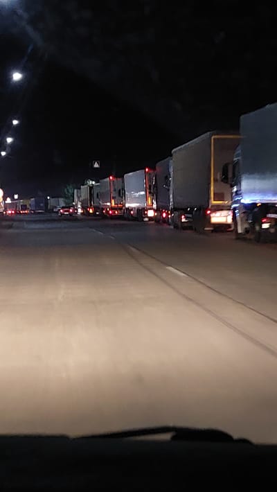 This media file depicts a traffic jam at night, likely in Дербісек ауылдық округі, Kazakhstan. The image is oriented vertically, with the vehicles and road appearing to run from bottom to top. A long line of trucks is visible, their brake lights glowing red in the darkness, indicating a standstill or slow-moving traffic. The road surface is a light-colored asphalt, with a distinct white line marking a lane. To the left of the road, the scene is dominated by the dark of night, with the vague shapes of trees or other roadside features visible. Some streetlights or distant lights are also visible, casting a soft glow. The overall impression is one of a tedious wait on a journey, with the focus on the repetitive nature of the truck taillights. There is no visible text in the image.