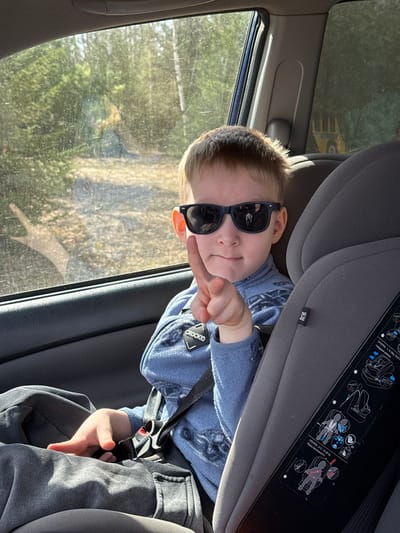 A young boy is pictured sitting in a gray car seat inside a vehicle, likely in Малая Уса, Russia. He is wearing dark sunglasses and a blue long-sleeved top, along with gray pants, and is secured by the car seat's harness. The boy looks playfully towards the camera, smirking, and extends his right hand with a "peace" or "victory" sign. Sunlight streams in from the window to his left, indicating a bright, sunny daytime setting. Outside the window, green foliage, trees, and hints of a structure or another vehicle are visible, suggesting an outdoor or rural environment. The car seat features several white safety labels with black text, including "CROCKID", "ГРУППА: 1/ 9-18kg ISOF", "Ремни безопасности", "ГРУППА: 2+3/15-36 kg", "BL1219AS0100-L", and "+якорный ремень", providing instructions for its use with different weight groups and belt types. The overall scene captures a candid, playful moment of a child traveling in a car.
