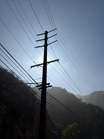 The image displays a utility pole in a mountainous, vegetated area of the Unincorporated Santa Monica Mountains, United States of America. The pole, positioned slightly left of center, stands vertically, featuring multiple cross-arms with visible insulators. A street light arm extends horizontally to the left from the lower section of the pole. Numerous utility lines are strung across the frame, connecting to the pole and extending diagonally across the bright, light blue sky. The sun is directly behind the upper half of the pole, causing it and the power lines to appear in stark silhouette. The background consists of dark, undulating mountains covered with dense vegetation, rendered in shadow and partially obscured by atmospheric haze. No individuals or specific human activity are present in the scene.