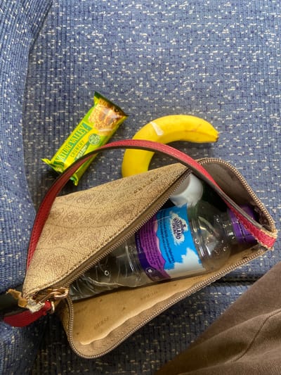 A still life scene is presented against a blue patterned surface, possibly a seat covering. A banana with a plaster on it, and a Nature Valley Oats & Honey Crunchy granola bar sit beside an open purse with a water bottle inside. The water bottle is labeled Highland Spring. The purse is patterned with the name "GUESS" printed in a repeating pattern. The purse is open, the contents are visible, and the purse appears worn, especially around the zipper and strap. The scene suggests the items are placed on a seat next to the purse. A person's brown clothing can be seen in the top right of the image. The picture was not taken in the City of Welwyn Hatfield.