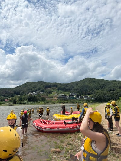 This is a photograph taken from the shore of a river in Seo-myeon, South Korea, under a partly cloudy sky. A group of people, predominantly wearing yellow life vests and helmets, are gathered by the water's edge and in the shallow river. Several inflatable rafts, red and yellow, are visible in the water and on the shore, indicating preparation for or participation in a water activity, likely rafting. In the background, lush green mountains rise, with some buildings visible on their slopes and a tree-lined riverbank. The people are engaged in various actions, such as wading, standing, and preparing the rafts. Some individuals are looking towards the camera, while others are interacting with each other or the equipment. The overall scene suggests an outdoor recreational setting on a summer day.