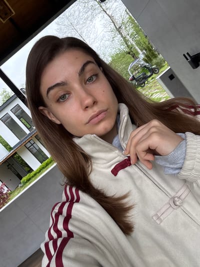 A young woman with light skin, brown hair, and green eyes is positioned in the foreground, looking directly at the camera. She is wearing a light-colored jacket with red stripes on the sleeves and a grey sweater underneath. Her hand is resting near the zipper of the jacket. The background shows a modern building with large windows, indicative of contemporary architecture. Outside the building, trees and greenery suggest a residential or suburban area. The scene is located in the city of Khala, Georgia. A motorcycle is visible through a window in the background.