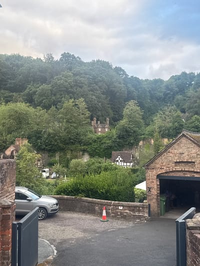 This image depicts a scene in Ironbridge, United Kingdom. The perspective is from a raised vantage point, looking down towards a wooded hillside and a street. In the foreground, a brick building with an arched opening, possibly a covered entryway or garage, is visible. A gray SUV is parked on the asphalt road in front of this building. Further down the road, a white traffic cone is placed on a brick wall.

The background features a densely wooded hillside with various buildings nestled amongst the trees. One building has a distinctive black and white timber-framed facade. The sky is overcast, suggesting it is daytime but potentially a cloudy or damp day. The overall atmosphere is somewhat subdued due to the weather and the green, natural surroundings. There is no discernible activity or event taking place; it appears to be a still, observational shot of the location. No visible text is present in the image.